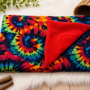 May include: A rectangular fleece blanket featuring a vibrant multi-colored tie-dye spiral pattern in shades of red, orange, yellow, green, and blue on a black background, with a solid bright red underside folded over at one corner.