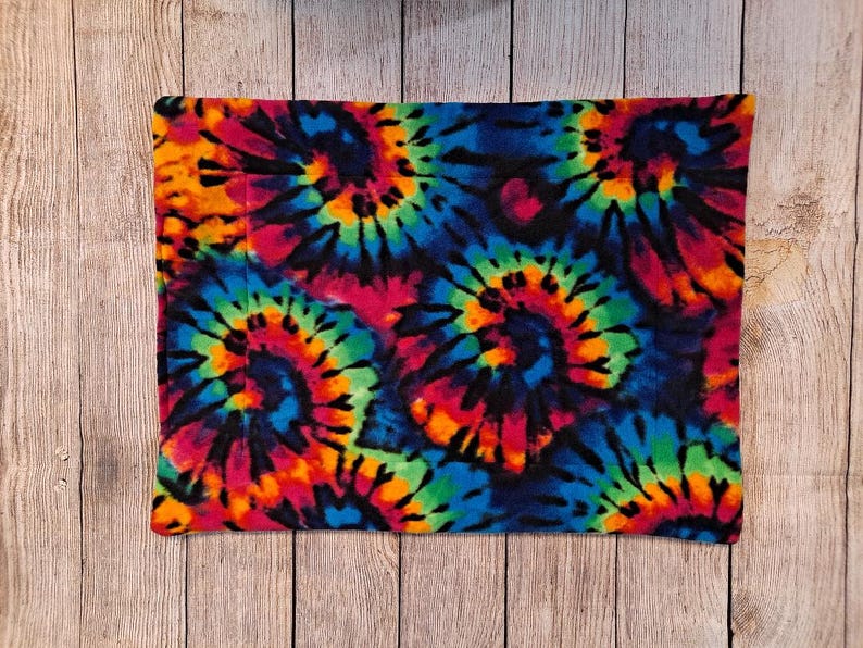 Może przedstawiać: Prostokątny kawałek materiału z żywym wzorem tie-dye, przedstawiający wirujące motywy w jasnych odcieniach niebieskiego, zielonego, ż&oacute;łtego, pomarańczowego i r&oacute;żowego na ciemnym tle. Tkanina posiada widoczne linie przeszyć biegnące pionowo.
