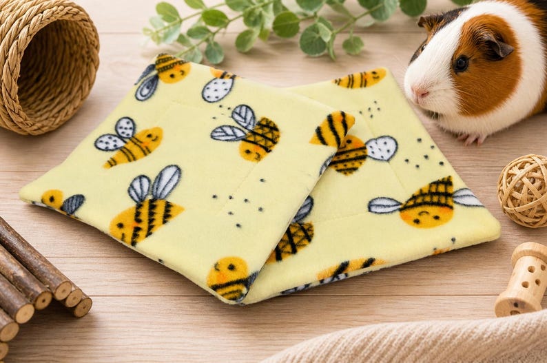 K&ouml;nnte beinhalten: Zwei quadratische, hellgelbe Fleece-Pads mit einem wiederkehrenden Muster aus Cartoon-Bienen mit schwarz-gelb gestreiften K&ouml;rpern und wei&szlig;en Fl&uuml;geln. Die weichen Stoffmatten liegen leicht &uuml;bereinander und zeigen das detaillierte Bienenmuster.