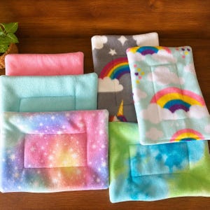 K&ouml;nnte beinhalten: Eine Sammlung rechteckiger Fleece-Haustiermatten in verschiedenen Mustern und Farben, darunter einfarbiges Pink, einfarbiges Hellblau, ein Regenbogen-Wolken-Druck, ein sternenreiches Galaxie-Muster und ein blau-gr&uuml;nes Batikmuster mit abgesteppten R&auml;ndern.