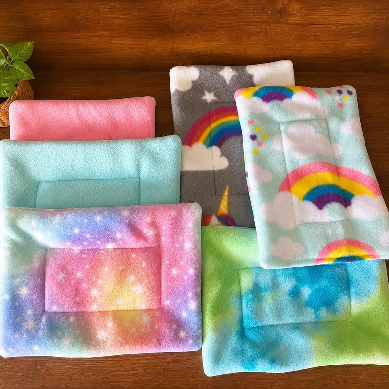 K&ouml;nnte beinhalten: Eine Sammlung rechteckiger Fleece-Haustiermatten in verschiedenen Mustern und Farben, darunter einfarbiges Pink, einfarbiges Hellblau, ein Regenbogen-Wolken-Druck, ein sternenreiches Galaxie-Muster und ein blau-gr&uuml;nes Batikmuster mit abgesteppten R&auml;ndern.