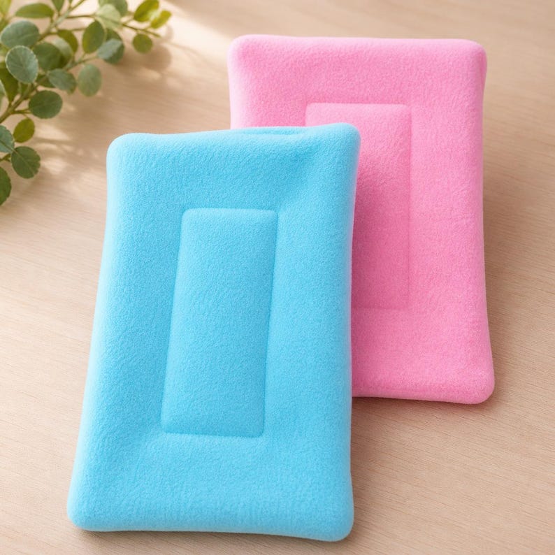 K&ouml;nnte beinhalten: Zwei rechteckige Kissen mit weicher Textur, eines in hellem Himmelblau und das andere in leuchtendem Pink. Jedes Kissen verf&uuml;gt &uuml;ber ein erhabenes, rechteckiges Mittelteil, das von einem glatten, pl&uuml;schigen Rand umgeben ist.