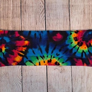 Może przedstawiać: Prostokątne owinięcie z tkaniny z żywym wzorem tie-dye z wirującymi kolorami niebieskim, r&oacute;żowym, zielonym, pomarańczowym i ż&oacute;łtym na czarnym tle. Materiał o miękkiej fakturze ma sp&oacute;jny nadruk na całej swojej powierzchni.