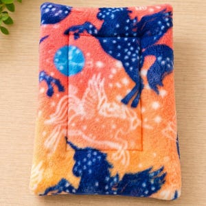 K&ouml;nnte beinhalten: Eine rechteckige Matte aus Pl&uuml;schstoff mit einem leuchtend orangefarbenen Hintergrund und einem wiederkehrenden Muster aus dunkelblauen Pferdesilhouetten, wei&szlig; umrandeten gefl&uuml;gelten Pferden und kleinen wei&szlig;en Sternen, erg&auml;nzt durch einen blauen Kreis.