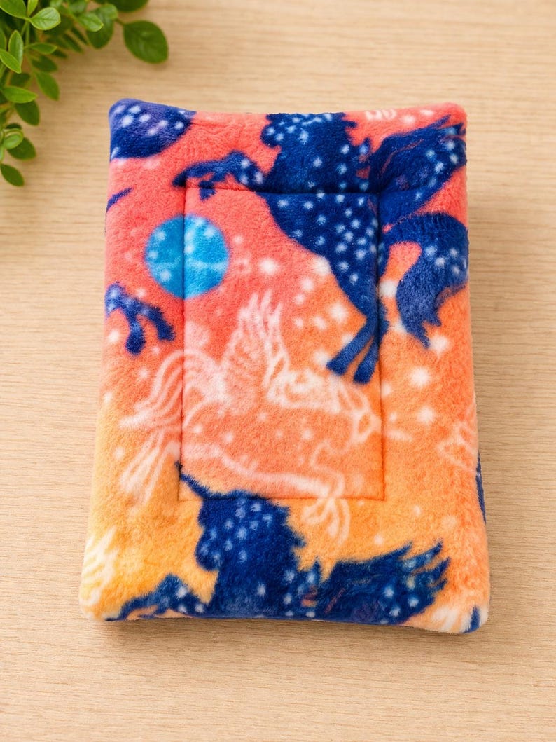 K&ouml;nnte beinhalten: Eine rechteckige Matte aus Pl&uuml;schstoff mit einem leuchtend orangefarbenen Hintergrund und einem wiederkehrenden Muster aus dunkelblauen Pferdesilhouetten, wei&szlig; umrandeten gefl&uuml;gelten Pferden und kleinen wei&szlig;en Sternen, erg&auml;nzt durch einen blauen Kreis.