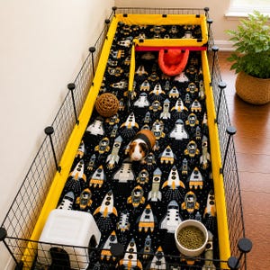 May include: A rectangular pet enclosure with black wire grid walls and a bright yellow fabric liner. The floor is covered in a black fabric featuring a repeating pattern of white and orange space rockets. A guinea pig, a white plastic hideout, a round woven ball, a red fabric bed, and a bowl of food pellets are inside.