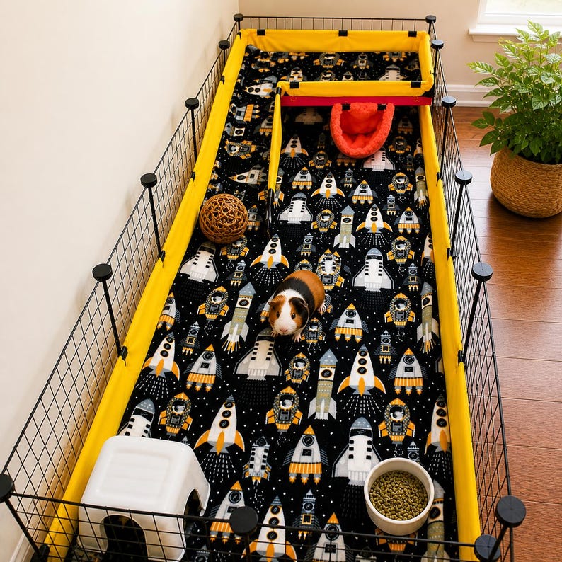 May include: A rectangular pet enclosure with black wire grid walls and a bright yellow fabric liner. The floor is covered in a black fabric featuring a repeating pattern of white and orange space rockets. A guinea pig, a white plastic hideout, a round woven ball, a red fabric bed, and a bowl of food pellets are inside.
