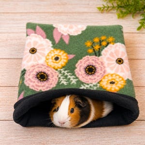 May include: A soft fabric tunnel with a dark green exterior featuring a floral pattern of pink and yellow flowers. A tricolor guinea pig with white, brown, and black fur is resting inside the opening, which is lined with solid black fabric.