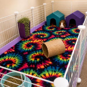 May include: A guinea pig enclosure featuring a tie-dye fleece liner with a spiral pattern in black, blue, green, yellow, and red. The cage includes a purple hay rack, a light blue food bowl, a tan cork tunnel, and two wooden houses.