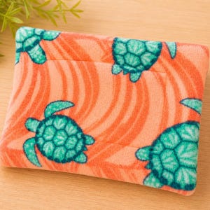 K&ouml;nnte beinhalten: Ein rechteckiges Etui aus weichem, flauschigem Stoff mit einem leuchtend korallenorangen Hintergrund, das ein sich wiederholendes Muster aus t&uuml;rkisfarbenen Meeresschildkr&ouml;ten und feinen, wellenf&ouml;rmigen, dunkelorangen Linien aufweist.