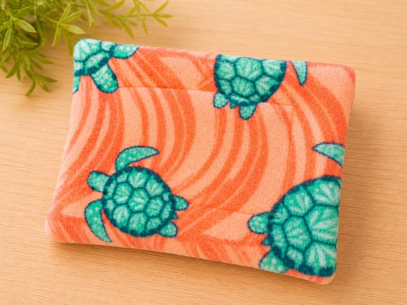 K&ouml;nnte beinhalten: Ein rechteckiges Etui aus weichem, flauschigem Stoff mit einem leuchtend korallenorangen Hintergrund, das ein sich wiederholendes Muster aus t&uuml;rkisfarbenen Meeresschildkr&ouml;ten und feinen, wellenf&ouml;rmigen, dunkelorangen Linien aufweist.