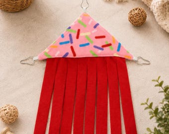 May include: A triangular fleece hammock featuring a pink top with colorful sprinkles and a red fringe bottom. The item includes three metal clips for suspension, with one at the top point and two at the base corners, designed for small animal enclosures.