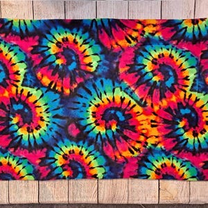 Może przedstawiać: Prostokątna poszewka na poduszkę z żywym wzorem tie-dye, przedstawiająca powtarzające się spiralne motywy w jasnych odcieniach neonowego r&oacute;żu, elektrycznego błękitu, słonecznego ż&oacute;łtego i limonkowego, na głębokim czarnym tle tkaniny.