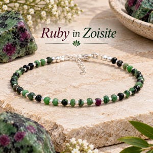 Op de afbeelding: Een kralenarmband met gefacetteerde robijn in zoisiet stenen afgewisseld met kleine zilverkleurige metalen kralen. De armband is voorzien van een zilverkleurige metalen verlengketting en een sluiting. De tekst Ruby in Zoisite is zichtbaar.