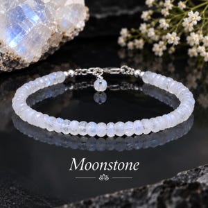 May include: A bracelet composed of small, faceted, translucent moonstone beads with a subtle blue iridescence. The bracelet features a silver-toned metal clasp and a small dangling moonstone charm. The text "Moonstone" is printed below the item.