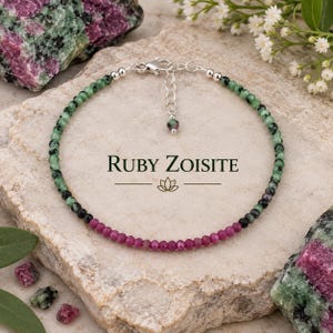 Puede incluir: Una pulsera de cuentas con piedras preciosas de rubí zoisita facetadas en verde y magenta, acentuada con cuentas de metal plateado, un cierre de mosquetón y una cadena ajustable. El texto "RUBY ZOISITE" está impreso con un icono de loto.