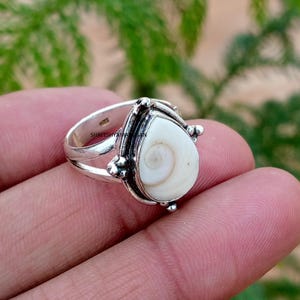 May include: A silver ring featuring a teardrop-shaped shiva eye shell inlay. The metal band has a split design and decorative beaded accents surrounding the central stone. The shell displays a natural spiral pattern in shades of white and cream.