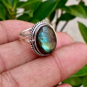 May include: A silver ring featuring an oval-shaped labradorite gemstone with blue and green iridescence. The stone is set in a polished silver bezel surrounded by a twisted rope-style metal border. The band has a split design with a smooth finish.