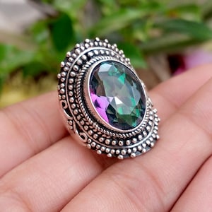 May include: A silver ring featuring a large, oval-cut mystic topaz gemstone with iridescent green and purple hues. The setting is crafted with intricate metalwork, including a beaded border and a twisted rope design surrounding the central stone.