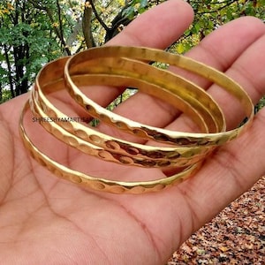 May include: Three gold-colored metal bangles featuring a hammered texture with rounded indentations along the outer surface. The text SHREESHYAMARTISAN is printed in black on the surface of the middle bangle, which has a polished finish.