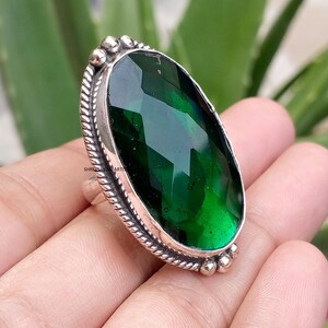 May include: A silver ring featuring a large, oval-shaped, faceted dark green gemstone. The setting includes a twisted rope-style border and small silver spheres at the top and bottom. The text SHREESHYAMARTIS is visible on the side of the metal.