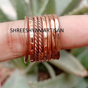May include: A set of seven copper rings featuring varied textures, including smooth, hammered, and twisted rope designs. The text SHREESHYAMARTISAN is overlaid across the centre of the rings, which are arranged side-by-side in a stacked formation.
