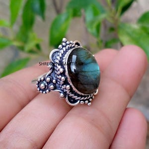 May include: A silver ring featuring an oval-shaped labradorite gemstone with blue and grey iridescence. The metal setting displays intricate beaded and twisted rope textures surrounding the stone. The text SHREE SHYAM ARTISAN is visible.