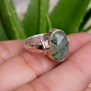 Può includere: Un anello in argento con una grande pietra verde ovale sfaccettata, caratterizzata da inclusioni scure simili ad aghi. La fascia presenta una finitura metallica lucida e un dettaglio decorativo a coste nel punto di incastonatura.