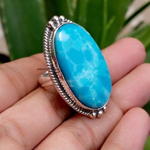 May include: A silver ring featuring a large, oval-shaped, bright turquoise stone with white marbling. The setting is framed by a twisted rope-like border and small silver beads at the top and bottom. The metal has a polished, slightly oxidised finish.