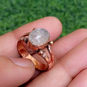 May include: A copper ring featuring an oval-shaped, translucent white gemstone set in a bezel. The band has a split design with small metallic spheres on either side of the stone. The text SHREESHYAMARTISAN is visible on the inner band surface.