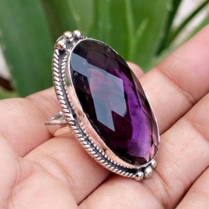 May include: A large oval ring featuring a faceted deep purple gemstone set in a polished silver frame. The metal setting displays a twisted rope texture along the border with small spherical accents at the top and bottom. The text SHREESHYAMARTISAN is visible on the side of the silver band.