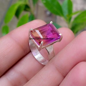 Pode incluir: Um anel de prata com uma grande pedra preciosa retangular facetada, apresentando um gradiente de cor do rosa profundo ao laranja. A pedra é fixada por quatro garras de prata. O texto SHREESHYAMARTISAN está impresso sobre a pedra.