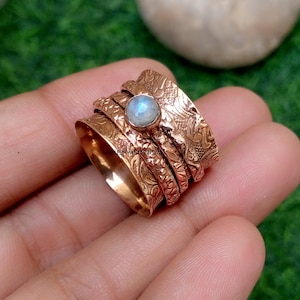 Op de afbeelding: Een koperen spinnerring met een reliëf bloemmotief over de brede band en een enkele ronde, iriserende maansteen cabochon in het midden gezet. Het metalen oppervlak heeft een gepolijste afwerking met ingewikkelde, verhoogde decoratieve details.