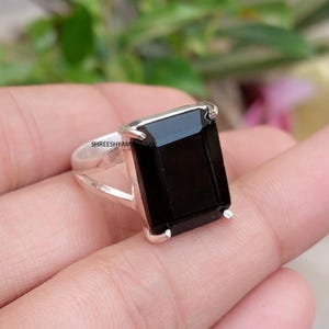 May include: A silver ring featuring a large, rectangular, faceted black gemstone held by four prongs. The band has a split design on the sides. The text SHREESHYAMARTISAN is visible on the side of the silver setting near the gemstone base.