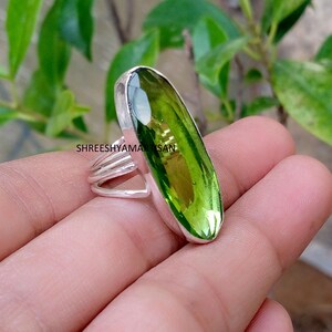 以下が含まれることがあります： 滑らかな金属製のベゼルにセットされた、大きく細長いファセットカットの楕円形の緑色の宝石が特徴のシルバーリング。バンドは3本の細い平行な銀線で構成され、ベースで収束しています。金属部分にはSHREESHYAMARTISANという文字が見えます。