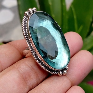 May include: A silver ring featuring a large, oval-shaped, faceted teal gemstone. The metal setting has a twisted rope-like border and small decorative spheres at the top and bottom. The band is plain silver and the stone has a translucent appearance.