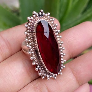 May include: A silver ring featuring a large, elongated, faceted deep red gemstone set in a decorative frame with a beaded, textured border. The metal setting displays a polished finish, and the text SHREESHYAMARTISAN is visible on the stone surface.