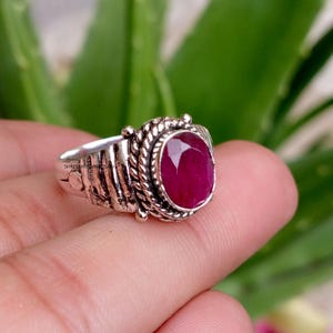Op de afbeelding: Een zilveren ring met een ovaalvormige, gefacetteerde donkerroze edelsteen in een zetting met een touwtextuur. De ringband heeft een brede, geoxideerde zilveren afwerking met verticale groeven aan de zijkanten en een klein gegraveerd logo.