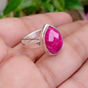 Puede incluir: Un anillo de plata de ley con una piedra preciosa de color rosa vibrante, tallada en forma de pera y engastada en un bisel de metal liso. La banda lleva grabado el texto 925 y SHREESHYAMARTISAN, indicando la pureza de la plata y la marca.