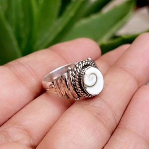 Può includere: Un anello in argento con un occhio di Shiva bianco centrale caratterizzato da un motivo a spirale naturale. L'incastonatura presenta un bordo a corda intrecciata e una fascia testurizzata a coste. Il metallo è lucido con zone scure.