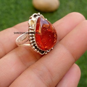Può includere: Un anello in argento sterling con una grande pietra d'ambra traslucida a forma di pera, di colore arancione intenso. L'incastonatura presenta un bordo in metallo perlato e il testo SHREESHYAMARTISAN è visibile sul lato della fascia in argento.