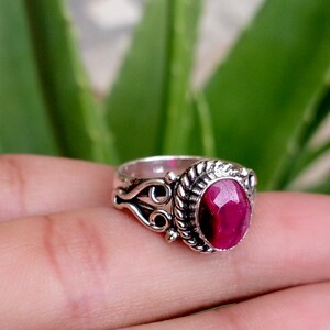 Peut inclure: Une bague en argent ornée d'une pierre précieuse rose foncé à facettes de forme ovale, sertie dans un bord en métal torsadé. L'anneau présente des détails complexes de volutes et de filigrane sur les côtés, encadrant la pierre centrale.