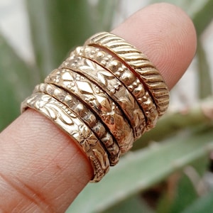 Pode incluir: Uma pilha de seis anéis de metal em tom dourado com várias texturas e padrões, incluindo estrias diagonais, pequenas esferas em relevo, formas geométricas de diamante, detalhes de contas e gravações florais, usados juntos num só dedo.