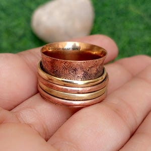Pode incluir: Um anel giratório de várias bandas com uma banda larga de cobre texturizado com o texto SHREESHYAMARTISAN gravado na borda interna, sobreposto com duas bandas de latão polido e duas bandas de cobre mais finas e lisas em design empilhado.