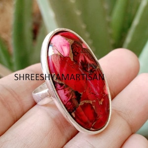 May include: An oval ring featuring a vibrant red stone with dark veining and metallic copper-colored inclusions, set in a polished silver-tone metal band. The text SHREESHYAMARTISAN is visible across the center of the stone surface.