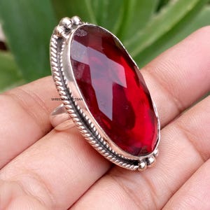 May include: A silver ring featuring a large, oval-shaped, faceted deep red gemstone. The setting is framed by a twisted rope-like metal border with small spherical accents at the top and bottom, showcasing a polished metallic finish throughout.
