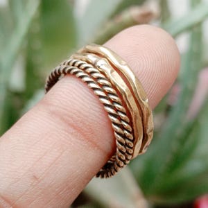 Peut inclure: Un ensemble de trois bagues empilées en métal doré. Le lot comprend deux bagues à la texture torsadée en corde et une bague à la surface lisse et martelée. Les bagues sont portées ensemble sur un doigt, mettant en valeur leur fini métallique.