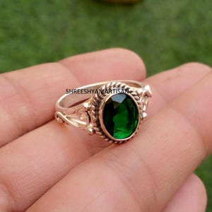May include: A silver ring featuring an oval-cut deep green gemstone set in a twisted rope-style bezel. The band displays intricate scrollwork and decorative metal detailing. The text SHREESHYAMARTISAN is visible on the metal surface.