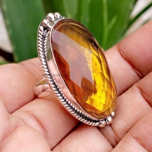 May include: A silver ring featuring a large, oval-shaped, faceted amber-coloured gemstone. The metal setting has a twisted rope-like border and small spherical accents at the top and bottom. The text SHREESHYAMARTISAN is visible on the side.