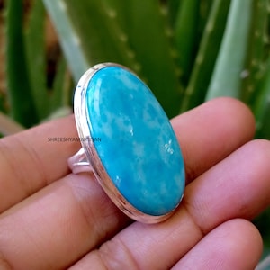 May include: A silver ring featuring a large, oval-shaped, bright turquoise blue gemstone with white mottled patterns. The stone is set in a smooth, polished silver bezel. The text SHREESHYAMARTISAN is visible on the side of the silver band.